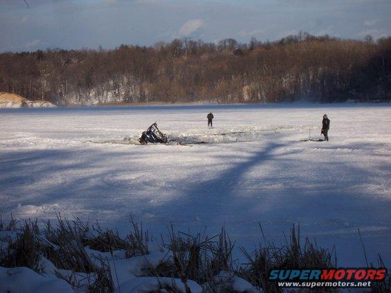 wreckage partially submerged in ice on a frozen lake, two people standing nearby