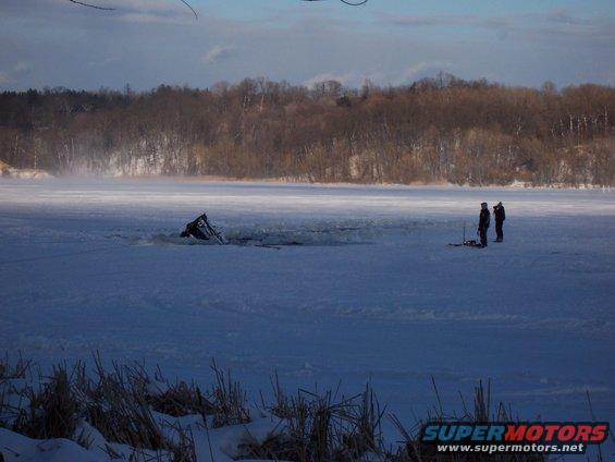Car partially submerged in ice on a frozen body of water, with two people standing nearby