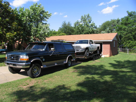 Green Ford Bronco SUV towing a white pickup truck on a trailer, parked on a driveway