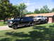 1995 Ford C-350 (Centurion Conversions) - A dark green Ford SUV tows a white pickup truck on a trailer in front of a brick house.