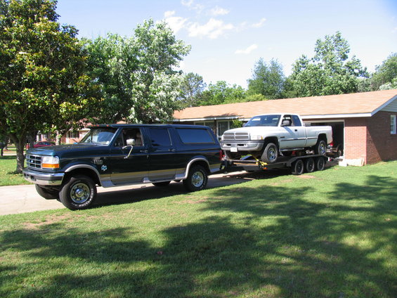 Dark green Ford SUV towing a white Dodge pickup truck on a triple-axle trailer in front of a house