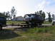 1995 Ford C-350 (Centurion Conversions) - A dark SUV tows a trailer carrying a black Ford Bronco past suburban houses.