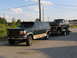 1995 Ford C-350 (Centurion Conversions) - Green Ford SUV towing a blue Ford Bronco on a trailer under a partly cloudy sky.