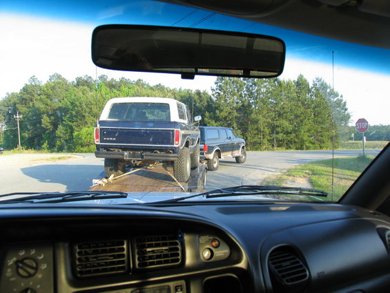 Rear view of blue Ford Bronco on trailer, lifted with large tires. Another dark SUV is visible on the road