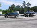 1995 Ford C-350 (Centurion Conversions) - A dark green Ford Excursion tows a blue Ford Bronco on a trailer, parked on a concrete lot with trees in the background.