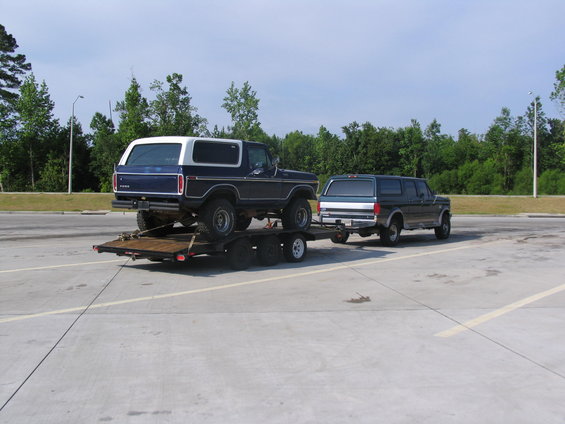 Blue and white Ford Bronco on a trailer, towed by a dark blue Ford SUV