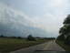 1995 Ford C-350 (Centurion Conversions) - A car travels down a rural road under a cloudy sky, with fields and trees lining the way.
