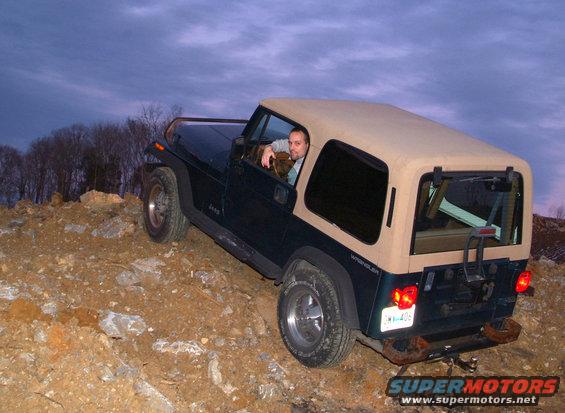 Jeep Wrangler with tan soft top driving on a dirt hill, driver visible