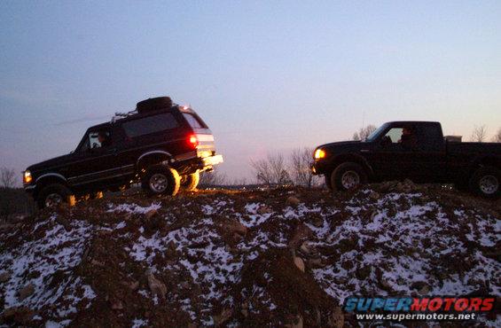 Black 1995 Ford Bronco with roof rack and spare tire on a snowy hill, next to a black Ford Ranger pickup truck