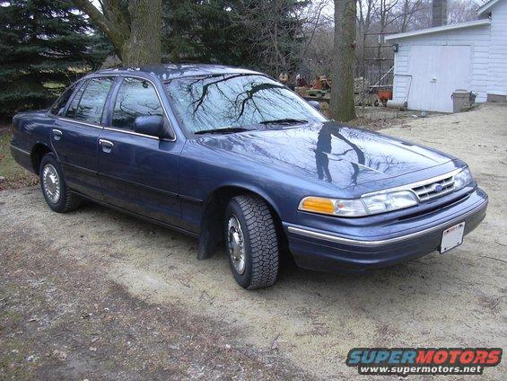 Blue sedan parked on dirt lot with trees and a white building in the background