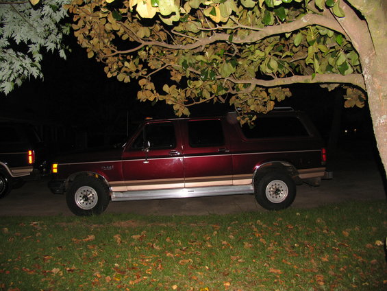 Maroon Ford Bronco parked at night under a tree. Another vehicle is partially visible to the left