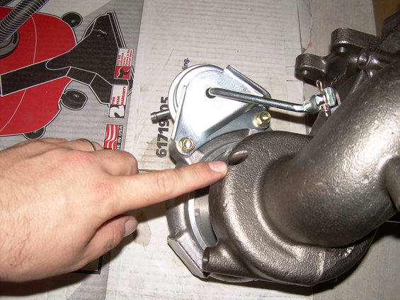 Close-up of a turbocharger with a finger pointing to a detail. Box with a red tool is in the background