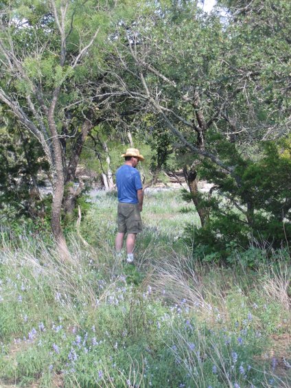Man wearing a straw hat and shorts stands in a grassy field with trees and wildflowers