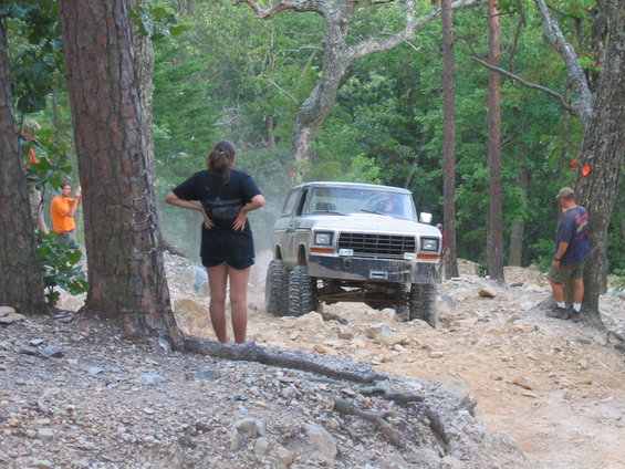 White SUV with large tires driving on a dirt trail with trees around. People are watching