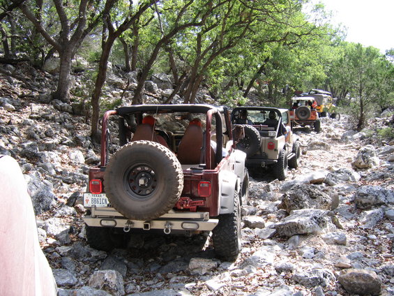 Several Jeeps driving single file on a rough, rocky path through a wooded area. The lead Jeep is dark red, followed by a tan one, then an orange one