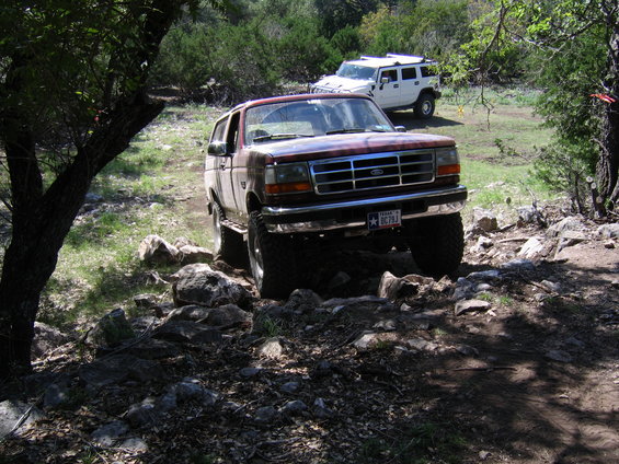 Red 1996 Ford Bronco driving over rocks on a trail, white Hummer in background