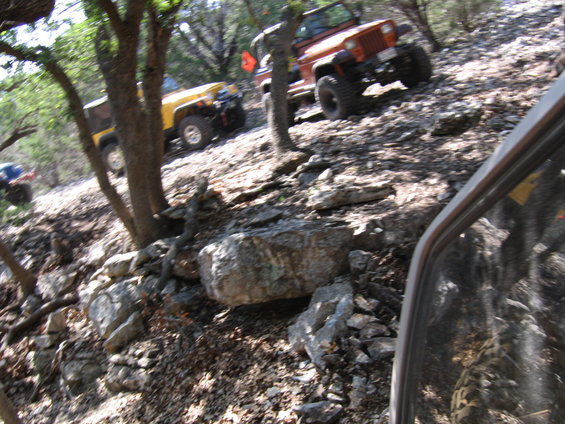 Orange and yellow Jeeps driving up a steep, rocky, wooded trail. View from inside another vehicle
