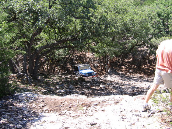 Blue Jeep Wrangler parked under trees on rocky terrain