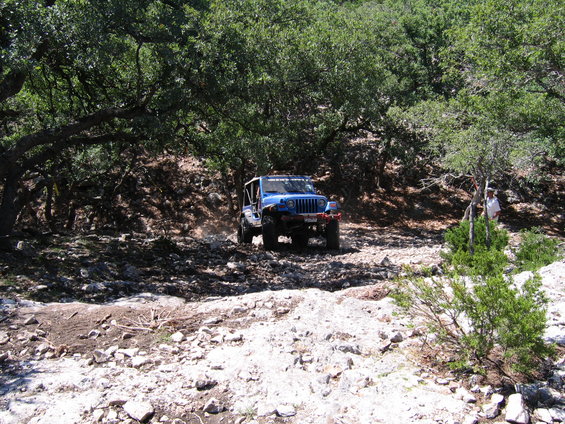 Blue Jeep Wrangler driving on a rocky, wooded trail. A person stands near some bushes on the right