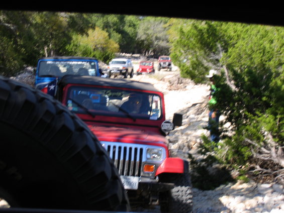 Several Jeeps driving in a line on a dirt and rock path through trees. A red Jeep is in the foreground