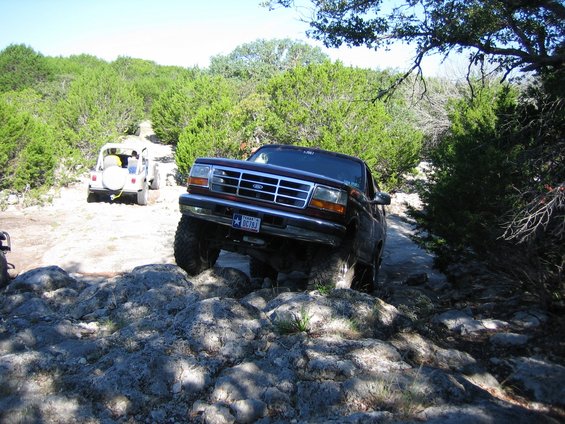 Dark red 1996 Ford Bronco navigating a rocky off-road trail with a white Jeep in the background