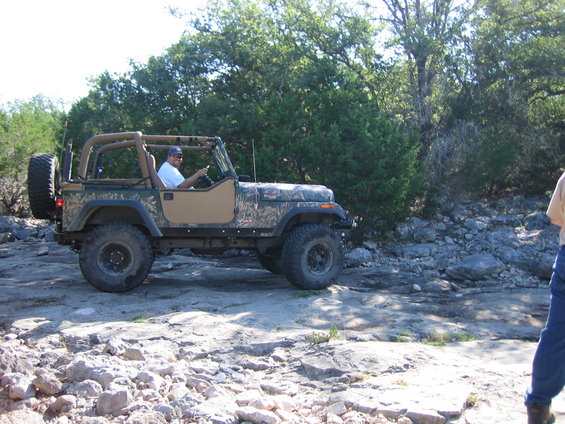 Camouflaged Jeep Wrangler driving on rocky terrain, tan soft top, spare tire on rear