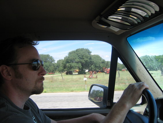 Interior view of a vehicle with a man driving, looking out at farm equipment in a field
