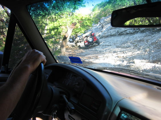 view from inside a vehicle driving on a rocky, wooded trail with another red vehicle ahead