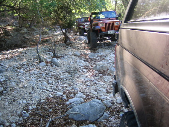 Orange Jeep Wrangler and another Jeep on a rocky, wooded trail, viewed from the side of a brown vehicle