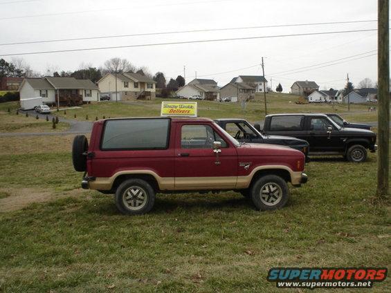Maroon Ford Bronco II parked on grass with two trucks behind it