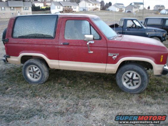 1986 Ford Bronco II, red with tan lower body trim, parked on grass. Another truck is visible behind it