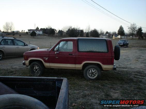 Red 1986 Ford Bronco II parked outside, spare tire mounted on rear door. Another SUV is parked to its left