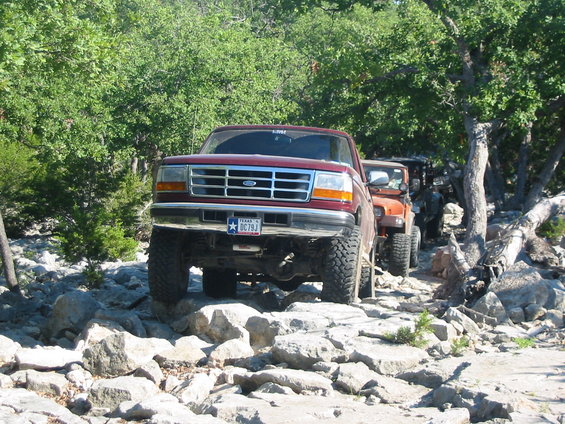 Red Ford F-150 pickup truck with lifted suspension and large tires, followed by an orange Jeep, driving on a rocky trail