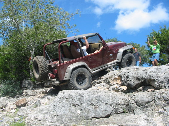 Maroon Jeep Wrangler TJ climbing over large rocks with two people inside and two people standing nearby