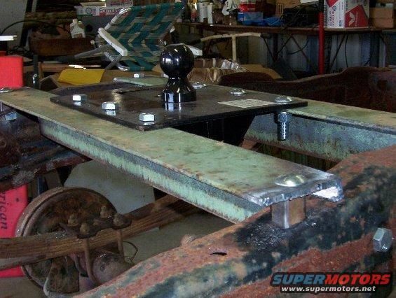Close-up of a fifth wheel hitch being bolted onto a rusty truck frame, with leaf springs and a wheel visible