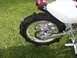 1900 Honda Big Red - Close-up of a motorcycle's rear wheel and exhaust system, set against a grassy background.