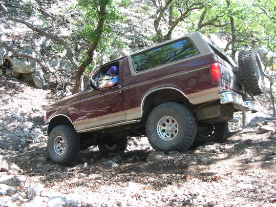 Maroon 1996 Ford Bronco with tan trim driving on a rocky, wooded trail. Spare tire mounted on rear