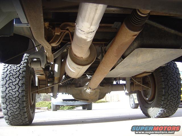 Underside view of a truck's rear axle, leaf springs, and exhaust system. Another vehicle is visible in the background