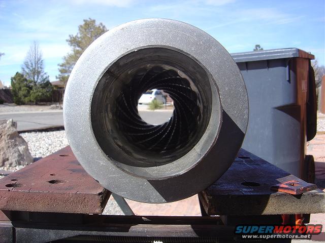Looking down the rifled bore of a large metal tube, with a trash can and a white vehicle visible in the background
