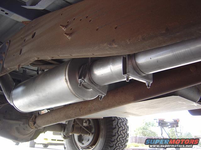 Underside of a vehicle showing a muffler and exhaust pipe, with visible rust on the frame