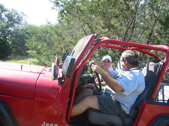 Man driving red Jeep Wrangler with young passenger in wooded area