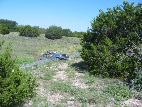 Blue SUV with its top removed stuck in a grassy ditch with purple flowers, viewed from a slightly elevated angle