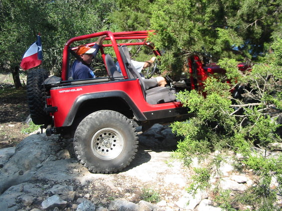 Red Jeep Wrangler with Texas flag on spare tire, driving on rocky terrain
