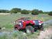 1996 Ford Bronco - A red Jeep Wrangler navigates a dirt path through a grassy, hilly landscape dotted with wildflowers under a clear blue sky.