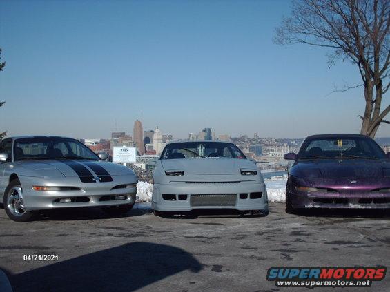 Three Ford Probes parked in front of a city skyline. One is silver with black stripes, one is grey, and one is purple