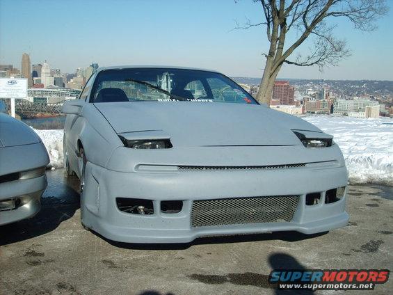 Gray car with pop-up headlights and custom body kit, front view, parked on pavement with snow in background