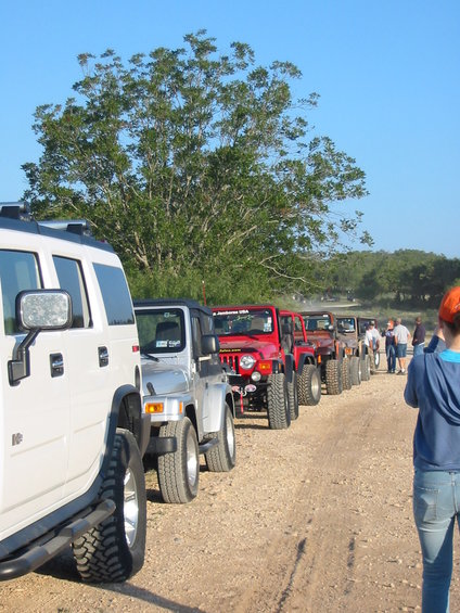 White SUV in foreground, followed by a line of Jeeps on a dirt road under a clear blue sky