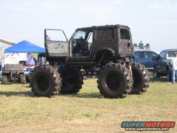 Mud-covered monster truck with huge tires, door open. Other vehicles and tents in background