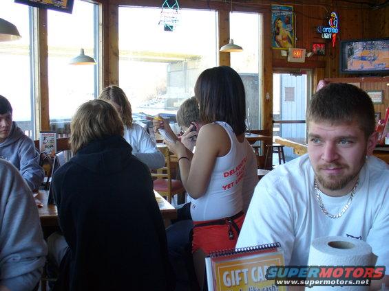 People sit at tables in a restaurant with windows showing an outdoor scene. A man in the foreground looks to the right
