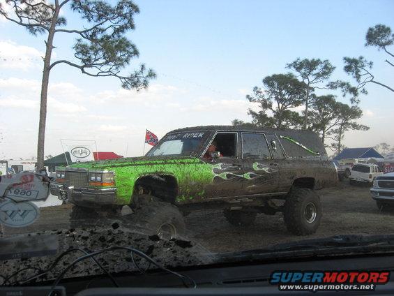 Mud-covered hearse with green flames and lifted suspension in a muddy field. Other vehicles and tents are visible in the background